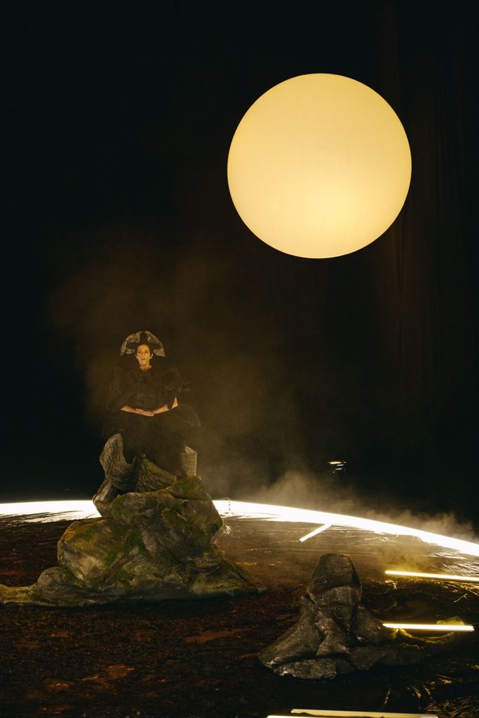A performer in black amid haze, illuminated by the sun and floor lights.