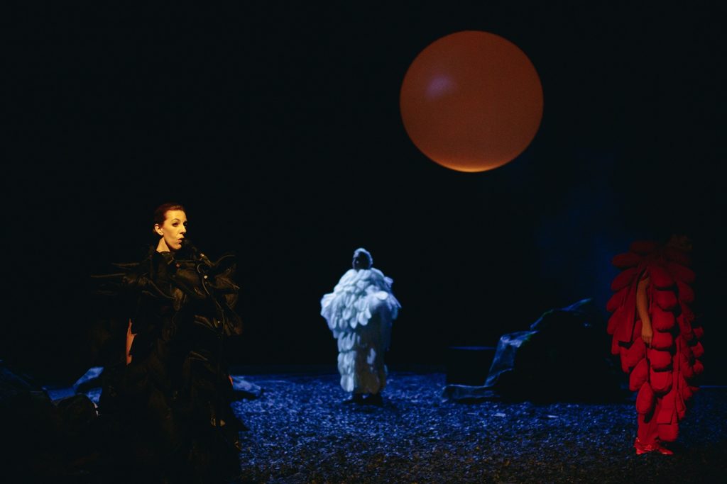 Three performers under a dim red sphere in a blue-lit landscape.