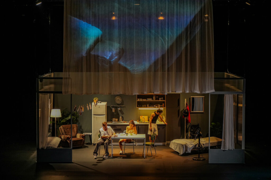 Three performers in a domestic interior beneath a projected image on a translucent curtain in Μεσοτοιχίες.