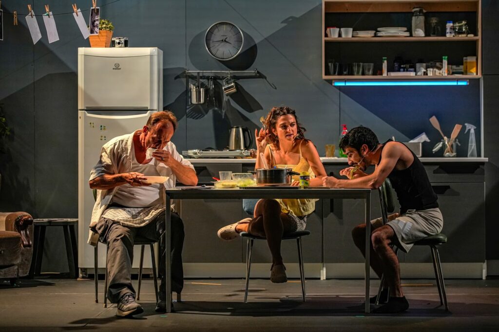 Three performers at a dining table in the domestic set of Μεσοτοιχίες.